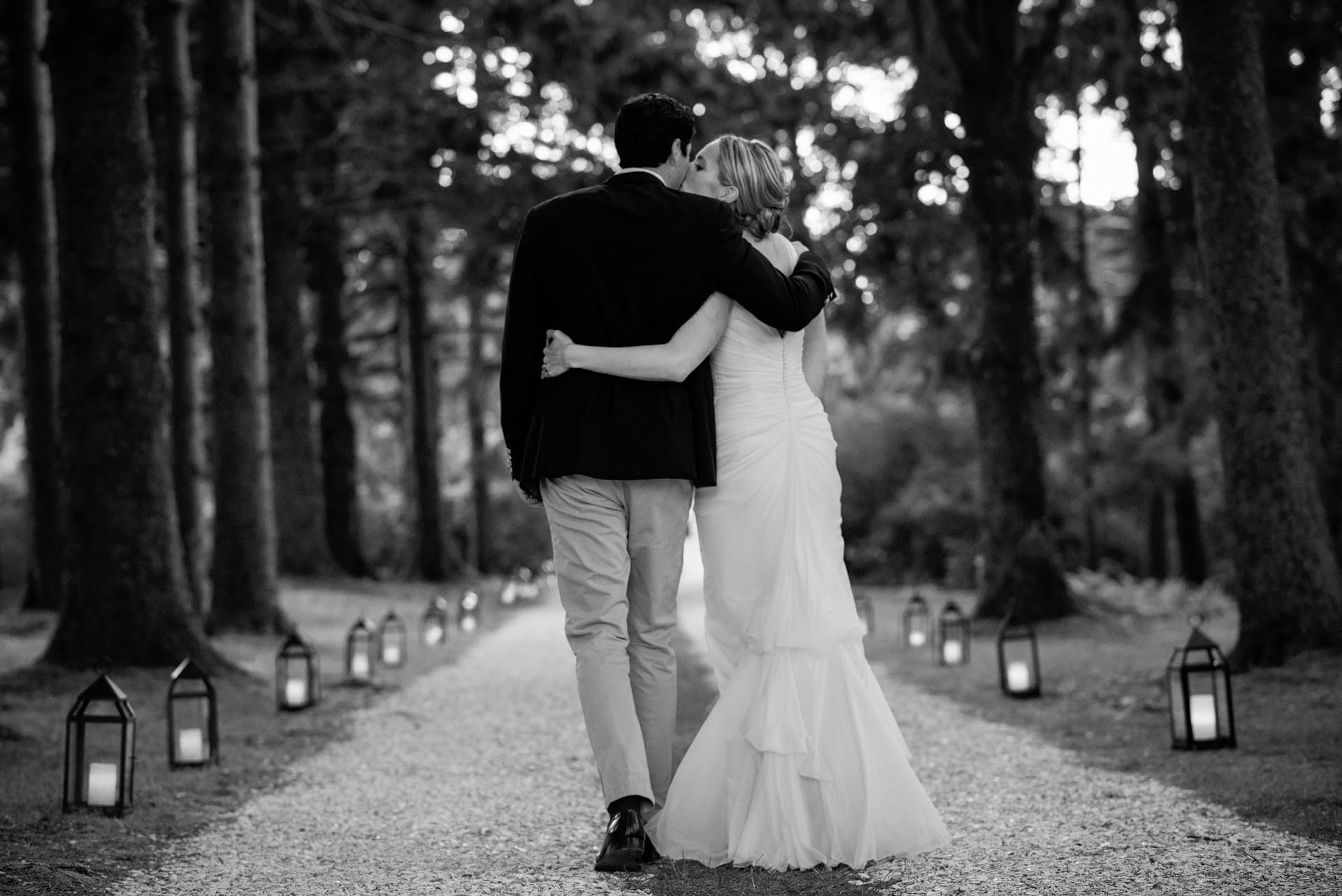 romantic black and white photo of couple walking down tree lined driveway with lanterns